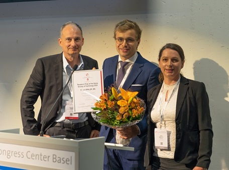 Drei Personen stehen vor einem Podium mit Blumen und feiern einen Gewinner eines der Wissenschafts-Awards von Amgen. Von links nach rechts: Prof Dr Richard Kobza, Dr Simon Kraler, Dr Anne Anstett